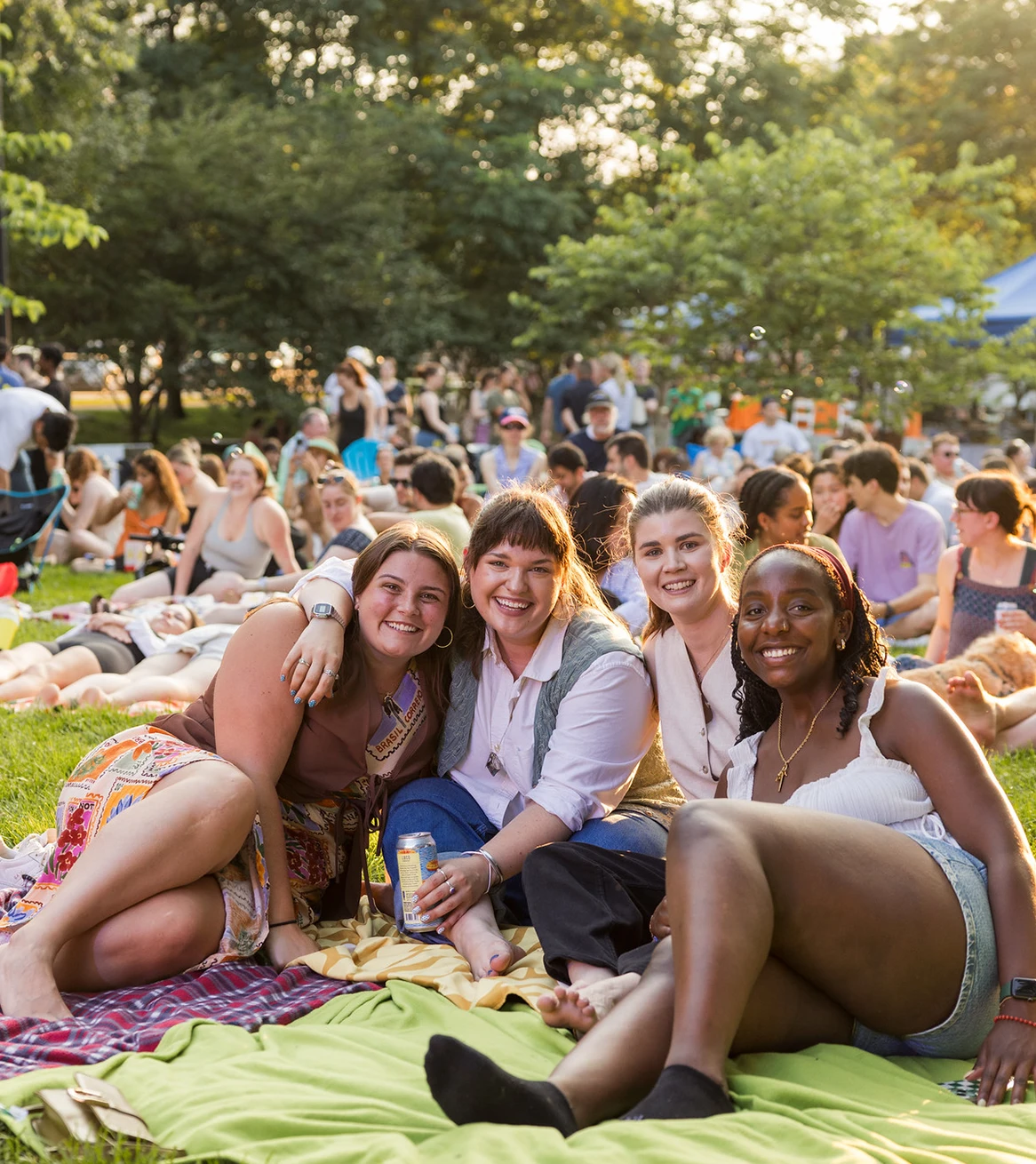Group of friends relaxing on picnic blanket in lively park during sunny outdoor community event