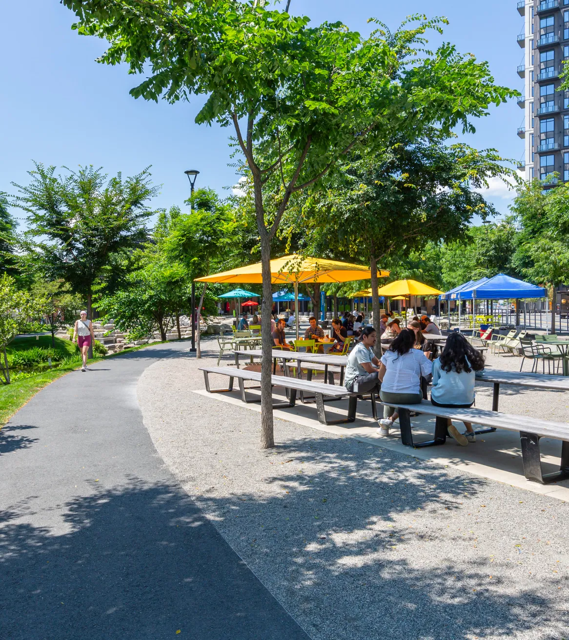 Outdoor park seating area with picnic tables and colorful umbrellas surrounded by trees and greenery
