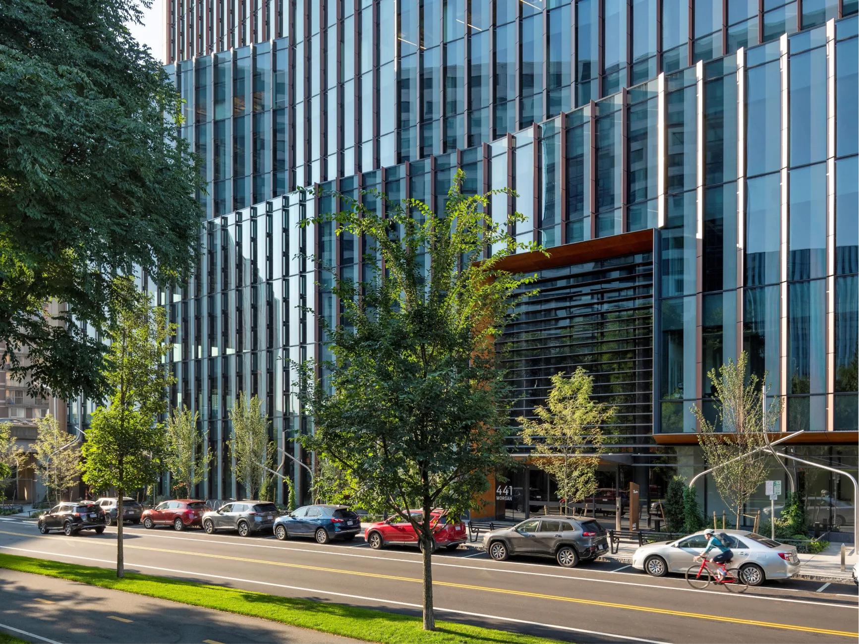 Modern glass office building facade along Cambridge street with trees parked cars and cyclist passing by